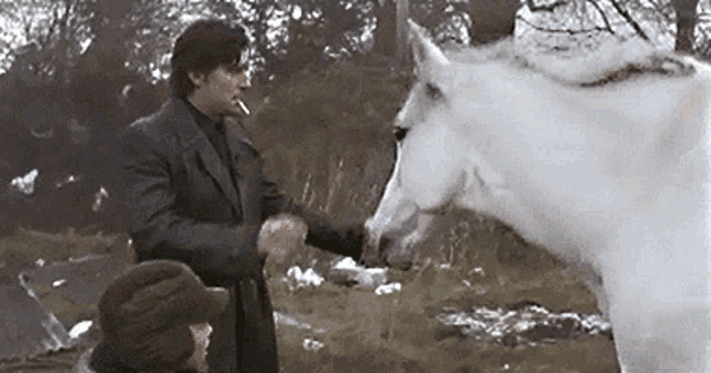 a man is smoking a cigarette while petting a white horse in a field .