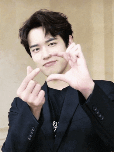 a young man in a black suit making a heart shape with his hands