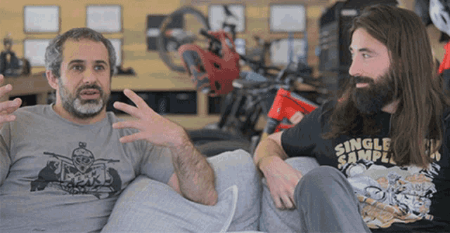 two men are sitting on a couch and one has a single sample shirt on
