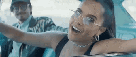 a woman wearing glasses is driving a car next to a man wearing a hat .