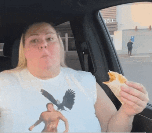a woman sitting in a car eating a sandwich with a bell sign in the background