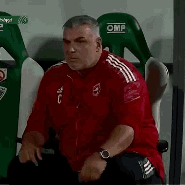 a man wearing a red adidas jacket is sitting in a green omp seat