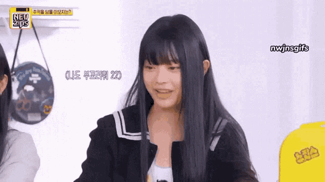 a girl with long black hair and bangs is smiling in front of a yellow sign that says nwjnsgifs