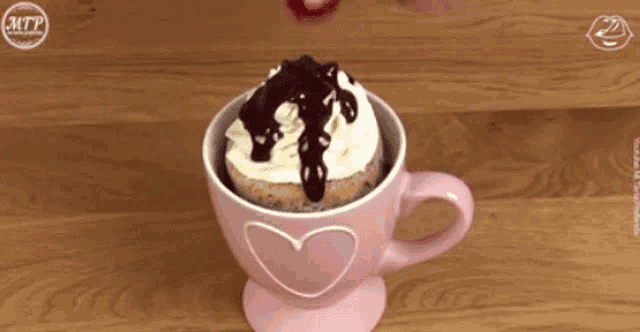 a pink cup with a heart on it has a cake in it with chocolate sauce and whipped cream