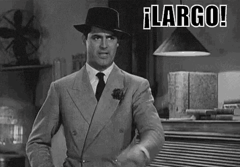 a man in a suit and hat is standing in a room with a fan and a lamp .