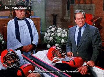 a man in a suit and tie is standing next to a coffin with a football player in it and a priest .