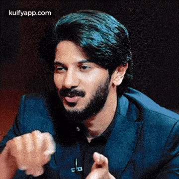 a man with a beard is wearing a blue suit and black shirt and is talking .
