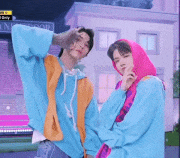 two men are standing next to each other in front of a building . one of the men is wearing a pink hoodie .