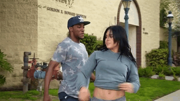 a man and a woman are dancing on a sidewalk in front of a building . the woman is taking off her shirt .