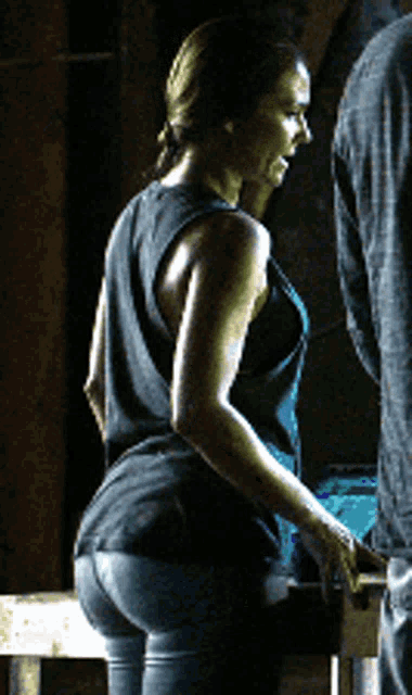 a woman in a black tank top and blue pants stands in a dark room