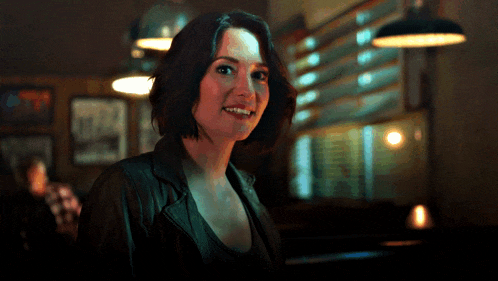 a woman in a black jacket smiles for the camera in a dark room