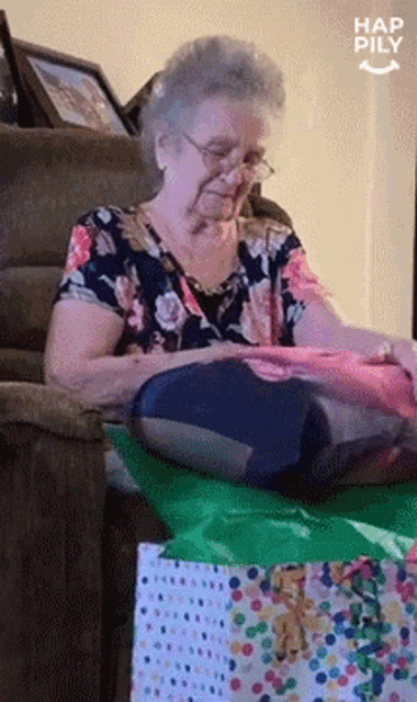 an elderly woman is sitting on a couch opening a gift bag with a happy pily logo in the corner