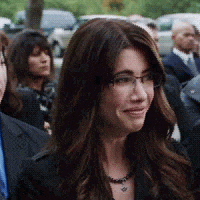 a woman wearing glasses and a necklace is standing in a crowd of people