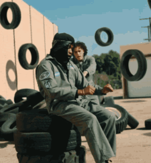 a man wearing a mask sits on a pile of tires with tires flying around him