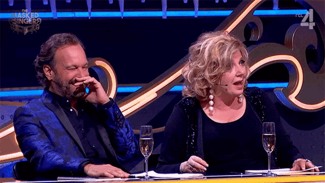 a man and a woman are sitting at a table with a masked singer logo on the wall behind them