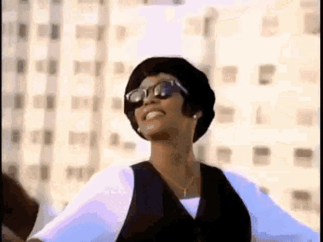a woman wearing sunglasses , a hat , and a vest is dancing in front of a building .