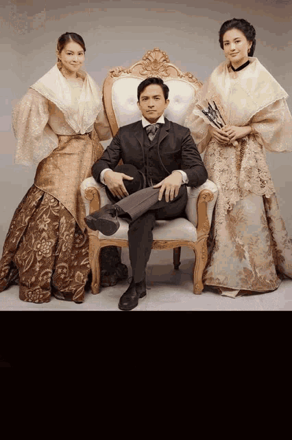 a man in a suit sits in a chair with two women standing next to him