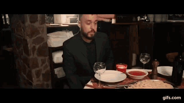 a man in a suit is sitting at a table with plates , bowls and glasses of wine .