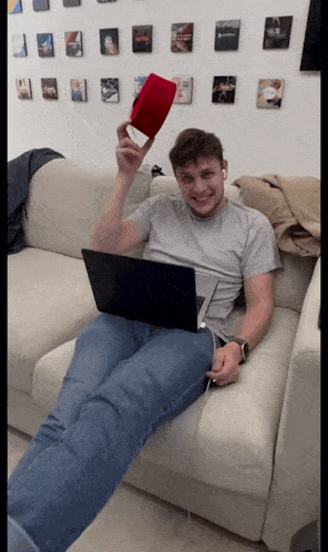 a man is sitting on a couch with a laptop and holding up a red object