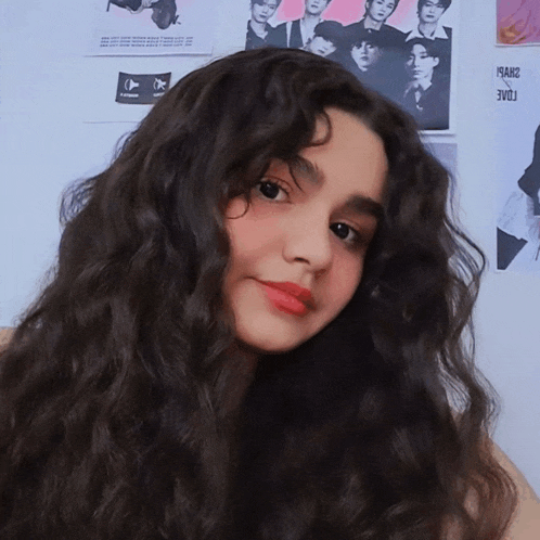 a girl with long curly hair is standing in front of a wall with posters of boys on it