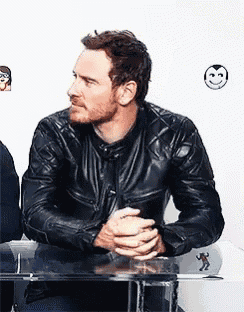 a man in a black leather jacket is sitting at a table with his hands folded and a smiley face behind him .