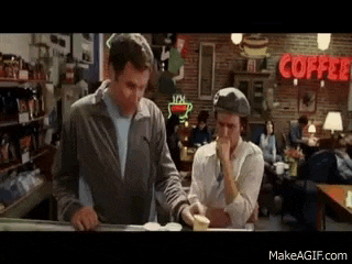two men are standing at a counter in a coffee shop with a coffee sign in the background .