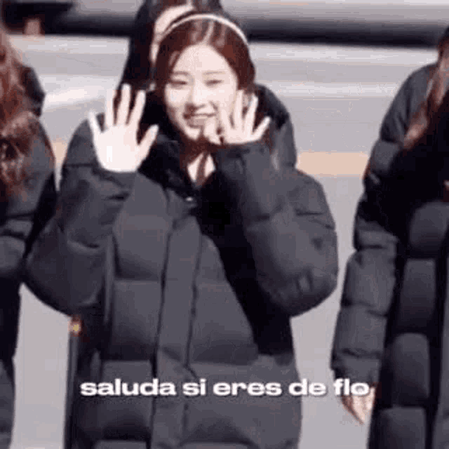 a woman in a black jacket is waving at the camera while standing next to a group of girls .