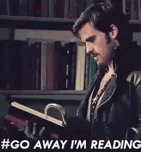 a man in a leather jacket is holding a book in front of a bookshelf and says " go away i 'm reading "