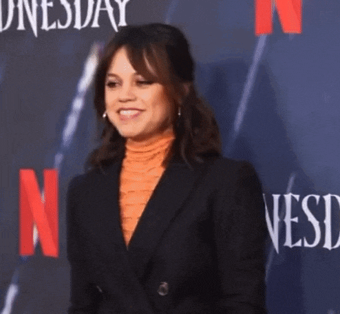 a woman in a black suit and orange turtleneck is standing in front of a netflix sign .
