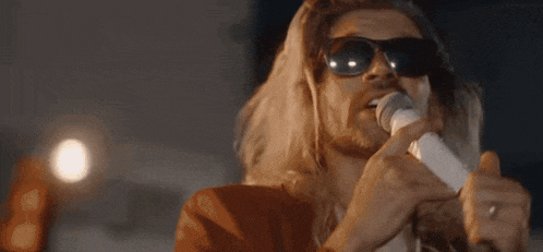 a man with long hair is singing into a microphone while wearing sunglasses .