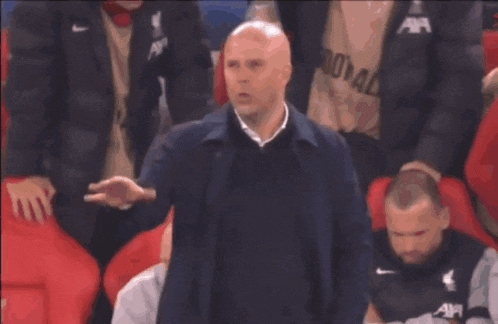 a bald man in a blue jacket stands in front of red chairs