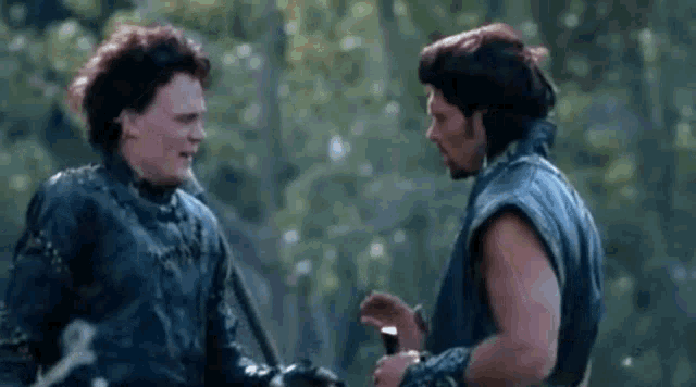two men are standing next to each other in a forest talking .