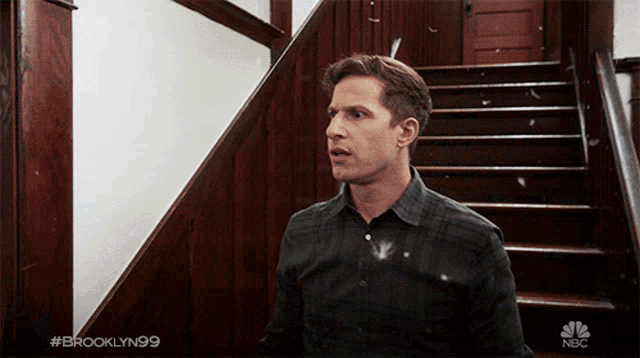 a man in a plaid shirt is standing in front of a set of stairs with #brooklyn99 on the bottom right