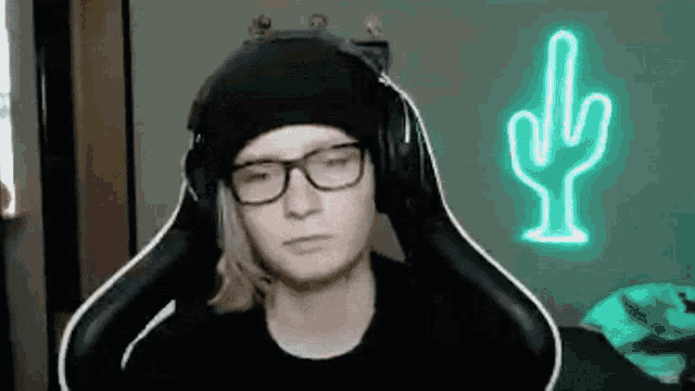 a man wearing headphones and glasses is sitting in a chair in front of a neon sign that says cactus .