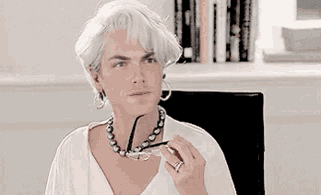 a man with white hair and a pearl necklace is sitting in a chair holding glasses .