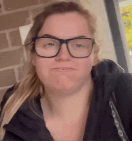 a woman wearing glasses and a black jacket makes a funny face .