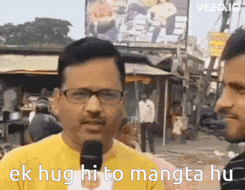 a man wearing glasses and a yellow shirt is talking into a microphone and says ek hug hi to mangta hu .
