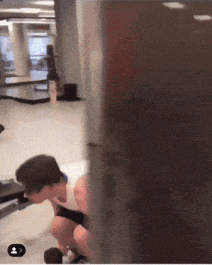a man squatting down in a gym with a person 's face visible in the corner