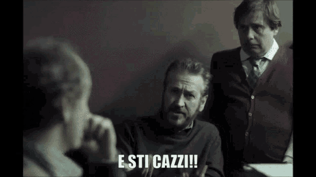 a man with a beard is sitting at a table with two other men and says e sti cazzi