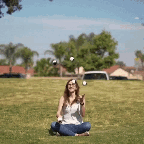 a woman is sitting on the grass juggling balls in a park .