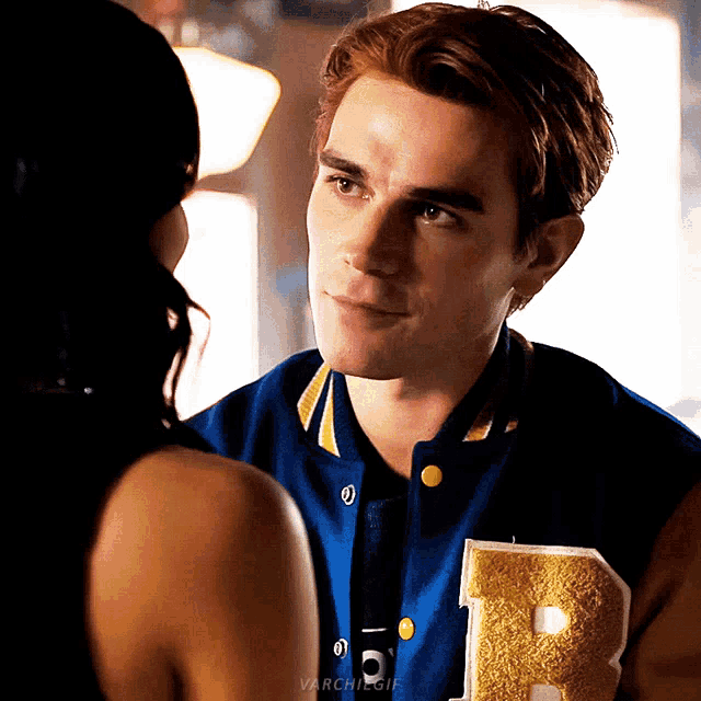 a man wearing a varsity jacket with the letter r on it looks at a woman