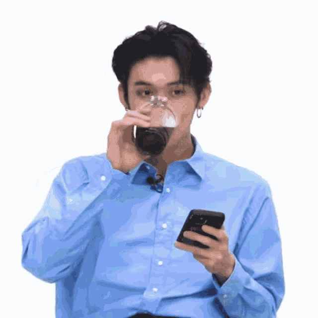 a man in a blue shirt is drinking a glass of soda while holding a cell phone .