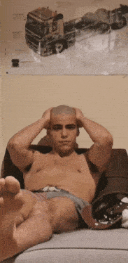 a shirtless man sits on a couch with his hands on his head in front of a poster of a truck