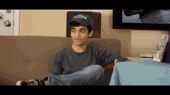 a young man is sitting on a couch with his legs crossed and wearing a hat that says california