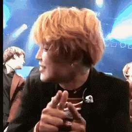 a man with blonde hair is sitting on a stage with his hands folded in front of his face .