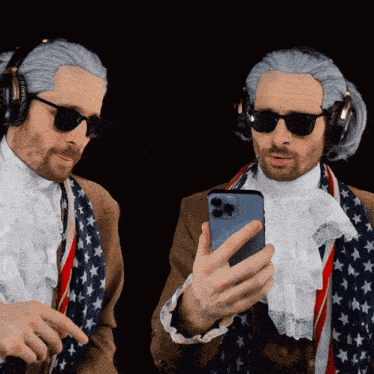 two men wearing wigs and headphones look at a cell phone