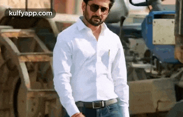 a man wearing a white shirt and sunglasses is standing in front of a blue tractor .