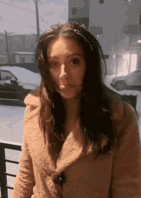 a woman in a tan coat is standing in front of a snowy street
