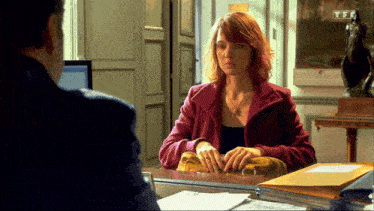 a woman in a red coat sits at a desk in front of a man with a tf logo on the screen