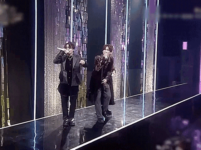 two men singing into microphones on a stage with purple lights behind them